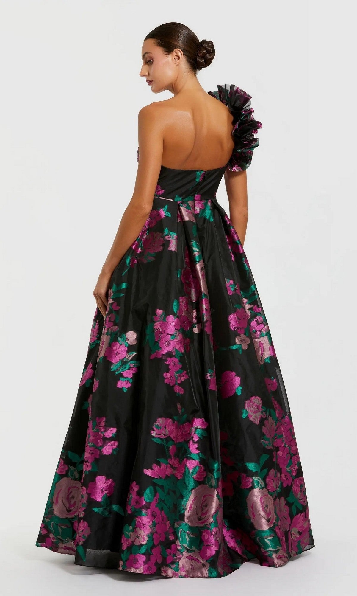 Woman wearing the Mac Duggal 11967 long formal dress—a black floral ballgown with pink and green flowers and a ruffled one-shoulder strap—posing against a plain light background, exuding classic elegance.